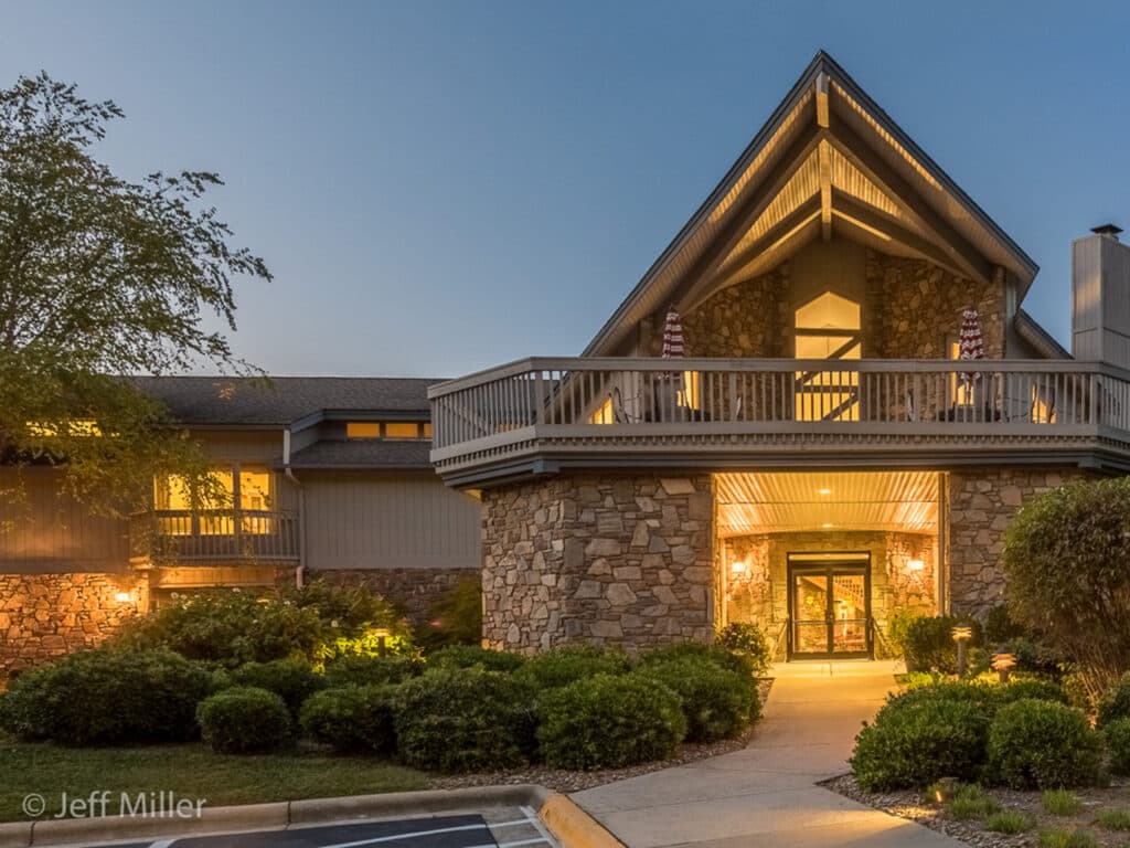 College Walk in Brevard is a large stone building with A-frame architecture, beautifully illuminated at dusk. Its well-lit entrance, surrounded by landscaped bushes, welcomes visitors. A charming balcony with railings adds to its appeal, along with a graceful tree on the left—perfect for independent living enthusiasts.