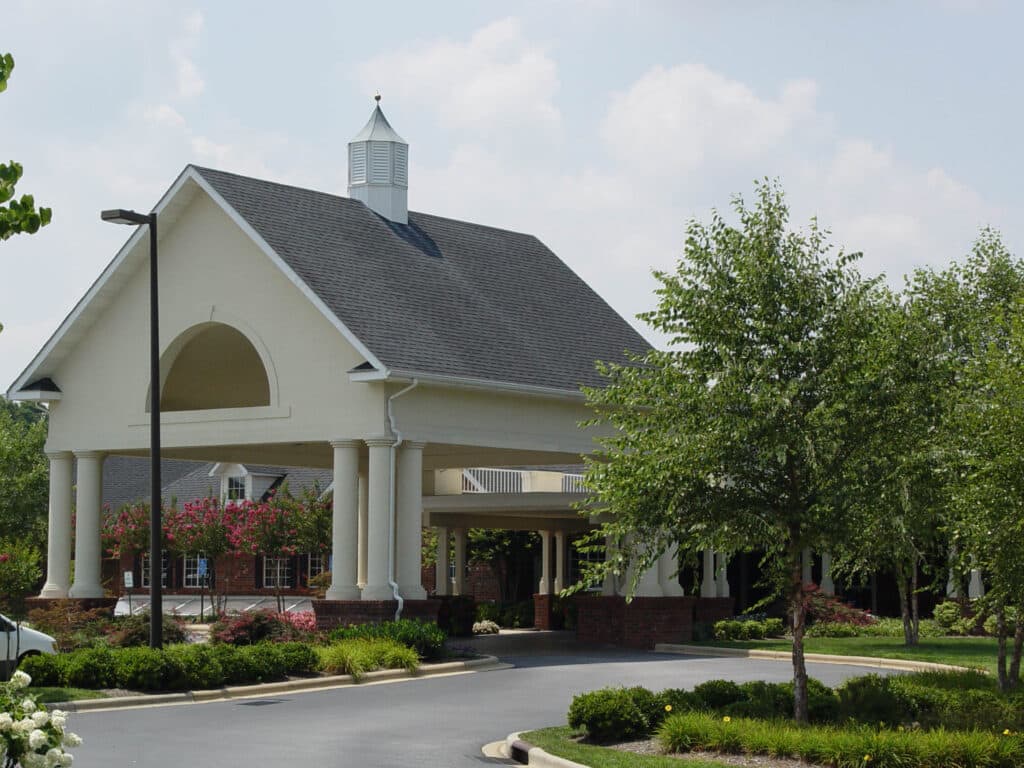 Lake Pointe Landing features a gabled white building with a cupola, perfect for senior living. Its surrounded by greenery and flowering shrubs, with tall columns supporting the grand entrance portico. A curved driveway welcomes visitors under the partly cloudy sky.