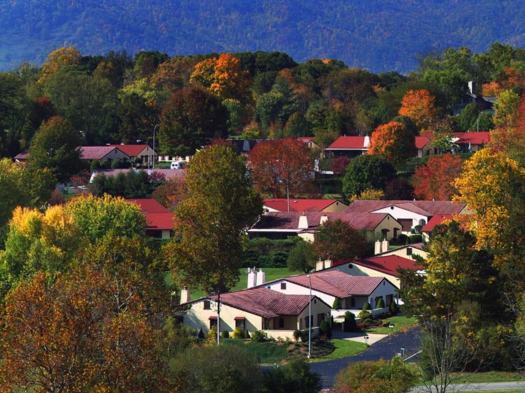 Discover Pisgah Valley in Asheville, where red-roofed houses are nestled amidst trees with vibrant autumn foliage. A forested hillside under a clear blue sky enhances this scenic view, perfectly suited for independent living.