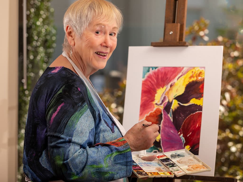 In a lively Asheville art studio, an older woman with short white hair from Ardenwoods smiles as she paints, embracing senior living. She holds a brush and palette filled with vibrant reds, yellows, and purples for her colorful abstract piece.