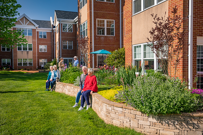 Residents relax on a garden retaining wall outside Tryon Estates, a spacious brick residence. They enjoy independent living among flowering plants under the shade of a turquoise umbrella. The tranquil scene encapsulates the Hendersonville lifestyle.